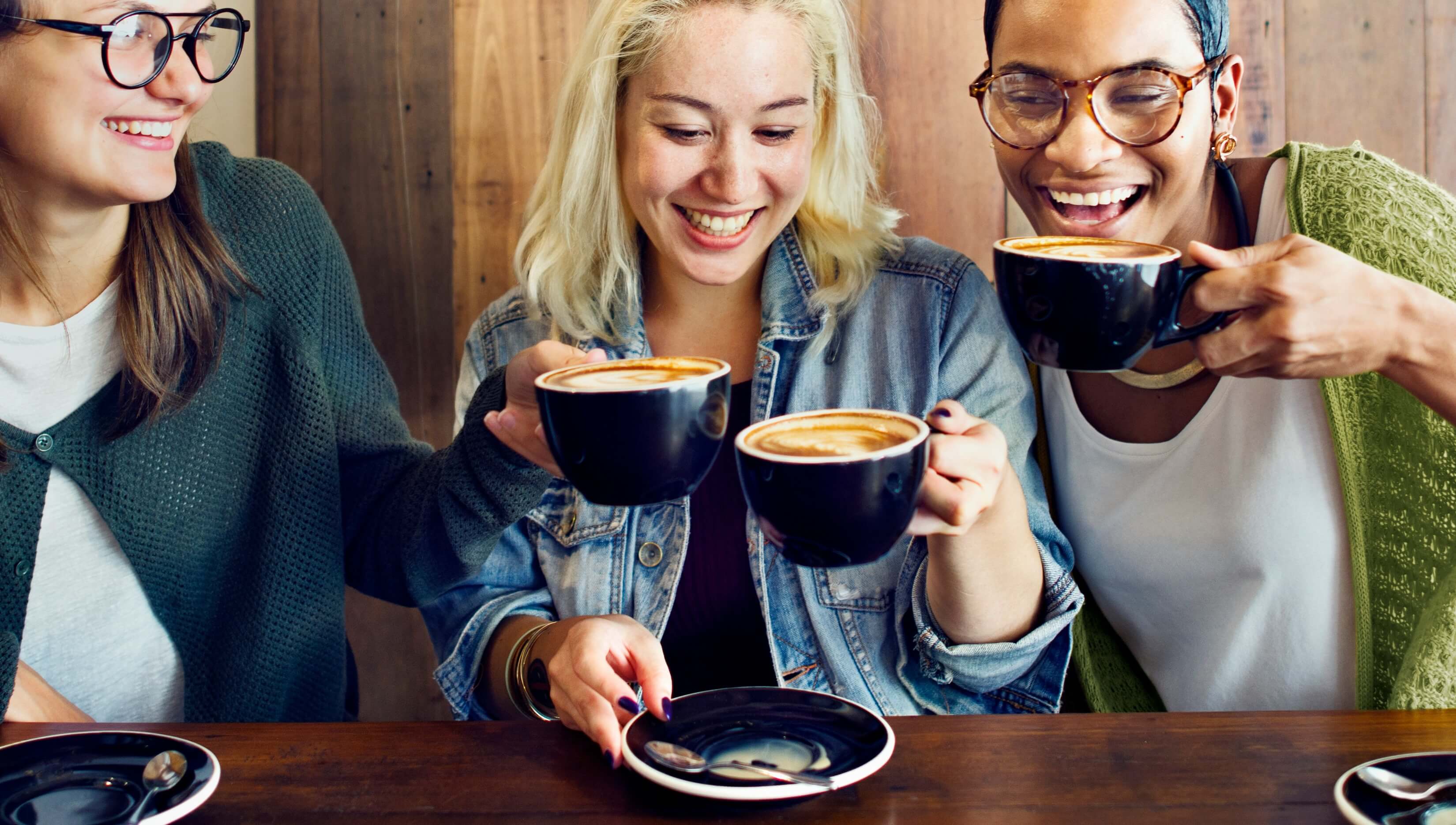 A young woman sitting across a small cafe table from two other women, mid-conversation over coffee, in a relaxed and warm setting.