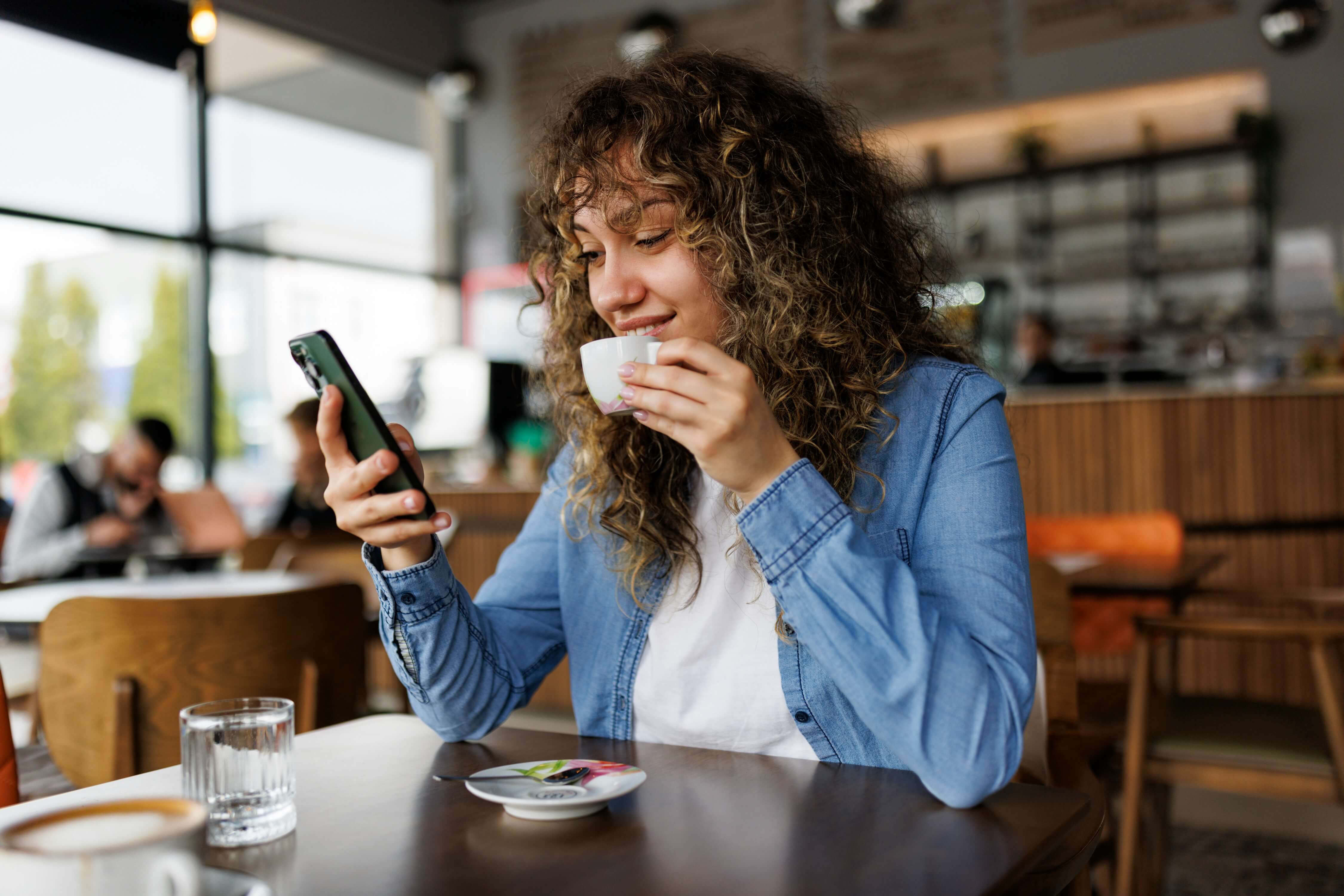A young woman scrolling Instagram on her phone at a sunny cafe table, reviewing her profile before sorority recruitment
