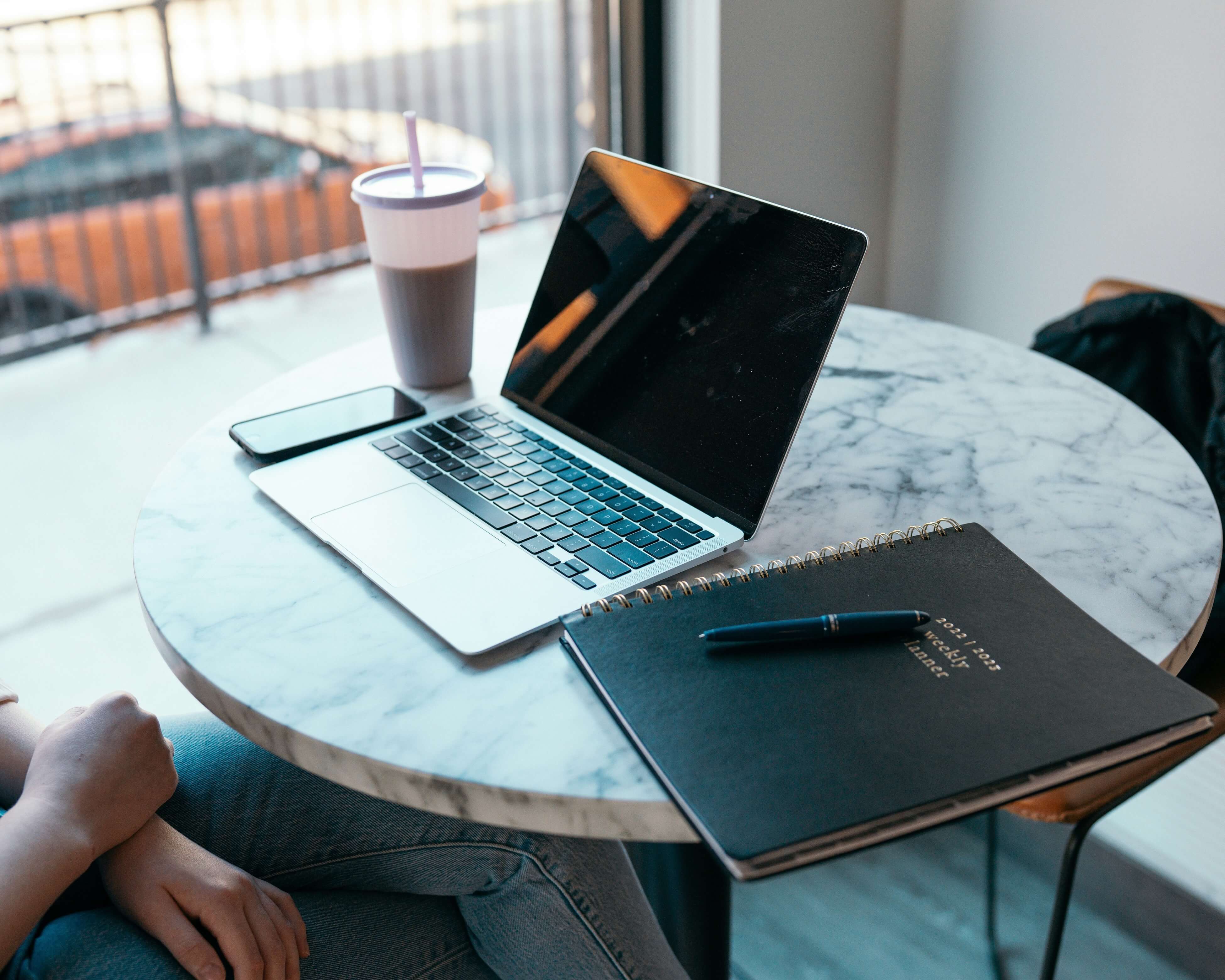 A sunlit desk with a laptop, notebook, and iced coffee, set up for summer sorority recruitment prep