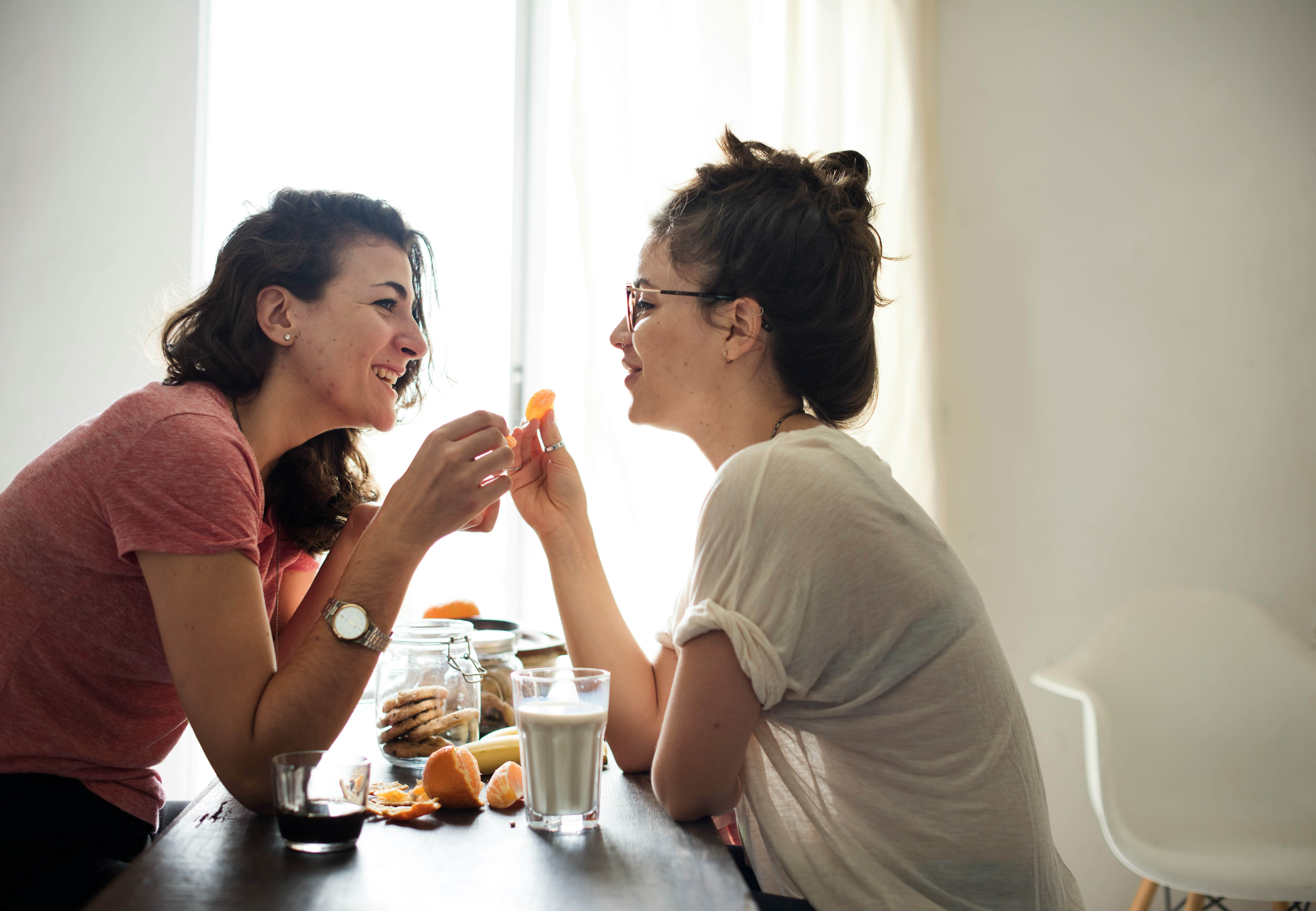 A mother and her college-age daughter splitting an orange at a kitchen table, mid-conversation, warm afternoon light.