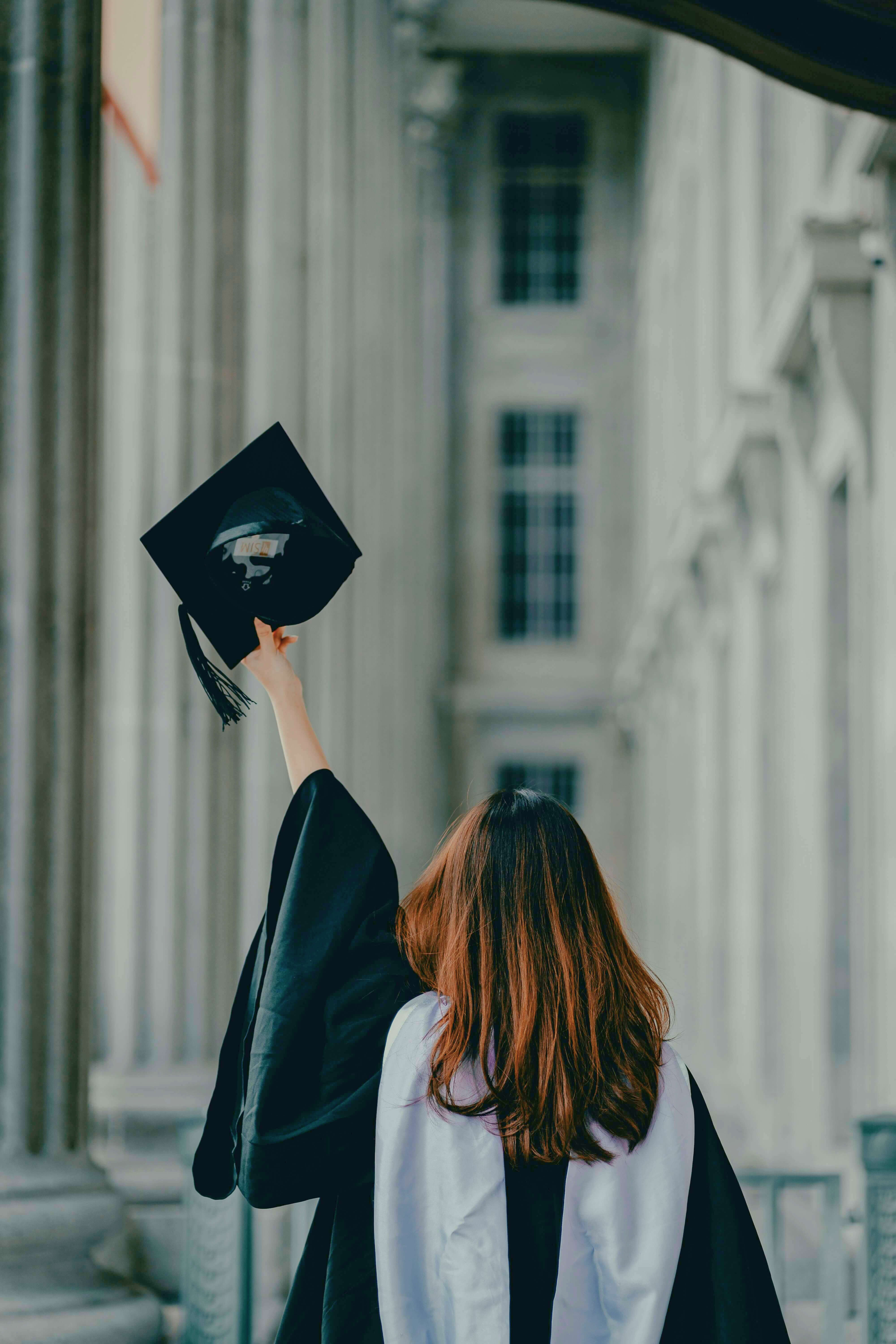 A young woman in graduation cap and gown, seen from behind, raising her cap in triumph outside a stone university building