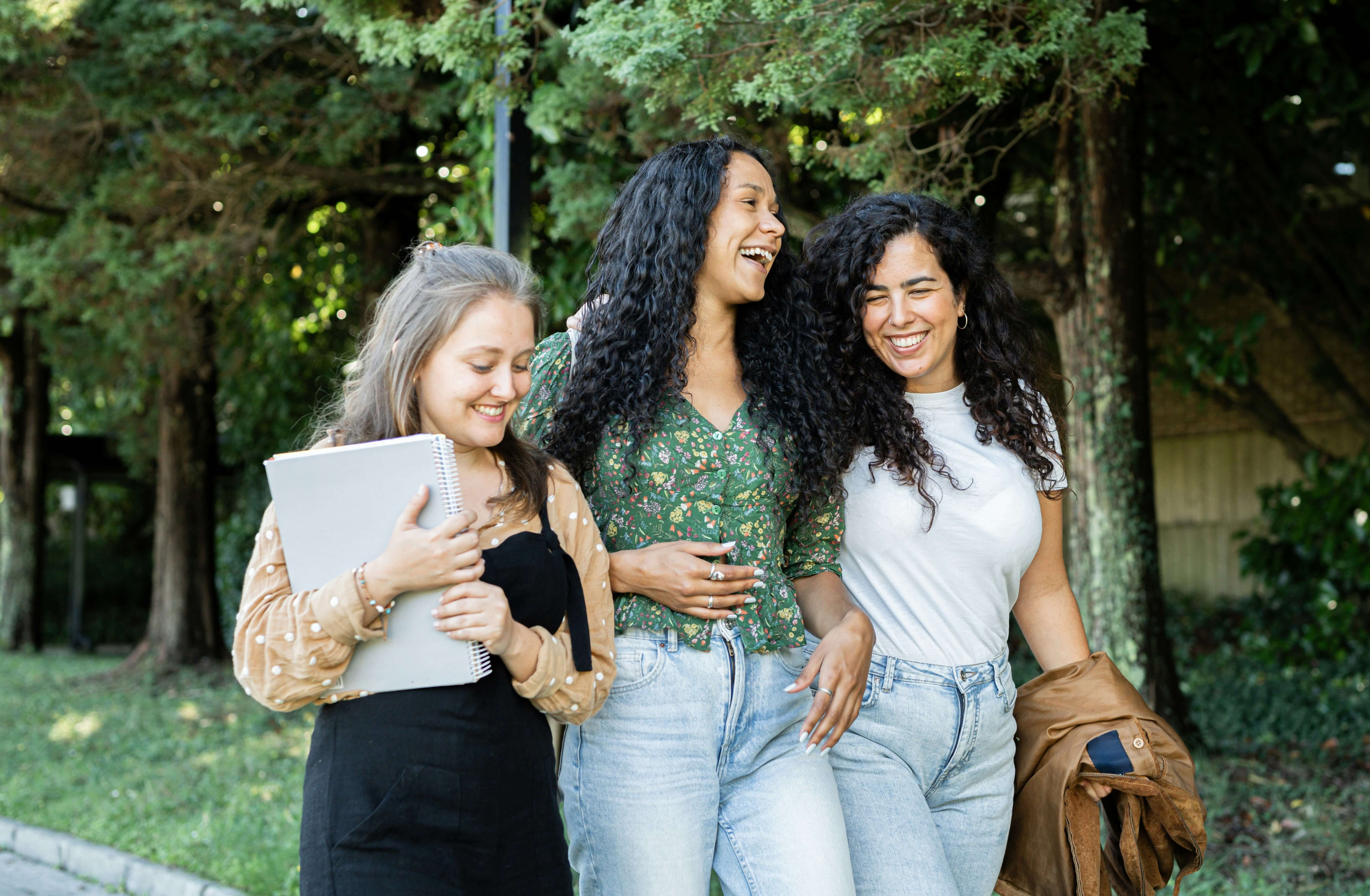 Three diverse college women walking and laughing together on a tree-lined campus path, one carrying a notebook