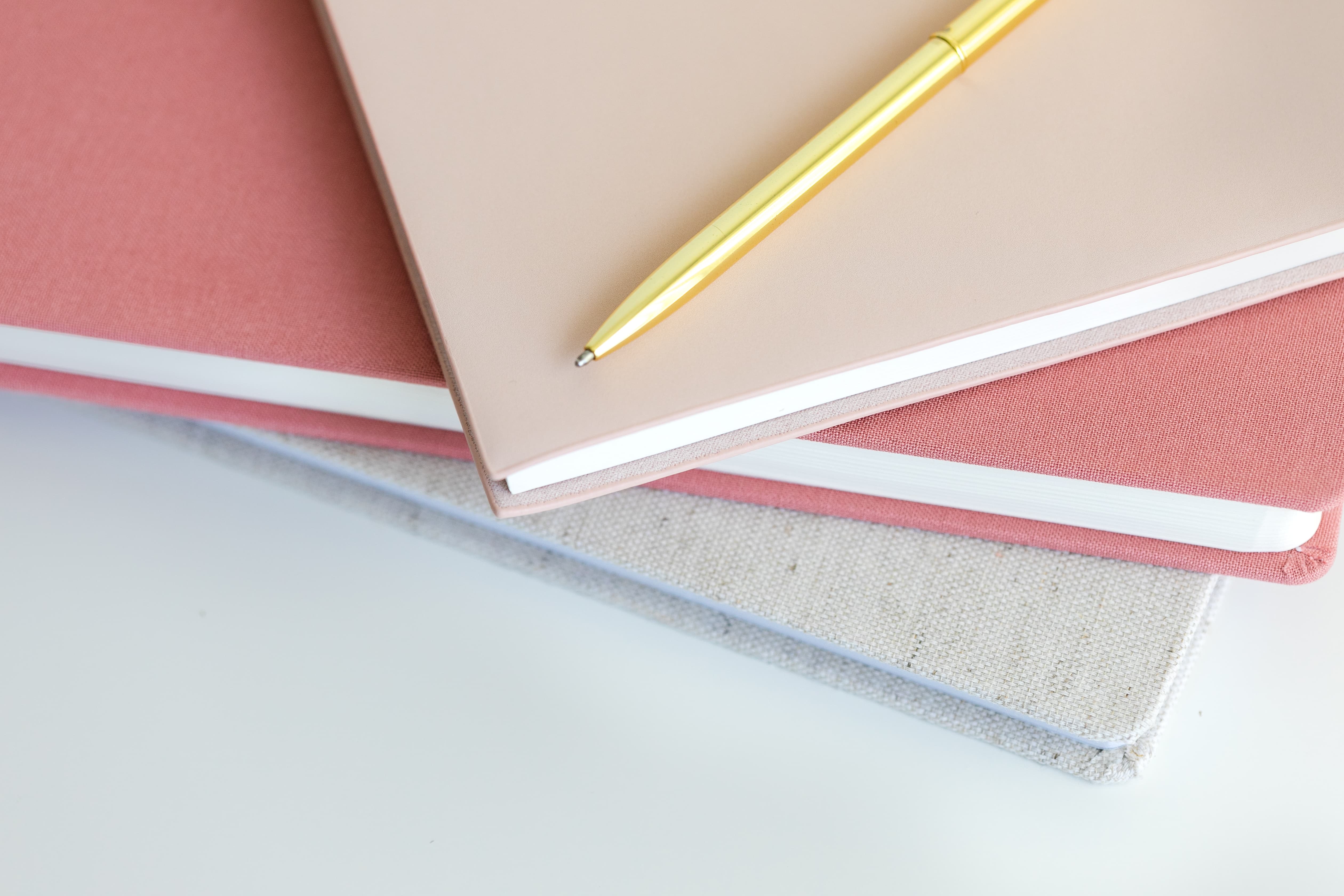Stack of three pink planning notebooks with a gold pen on a white marble surface