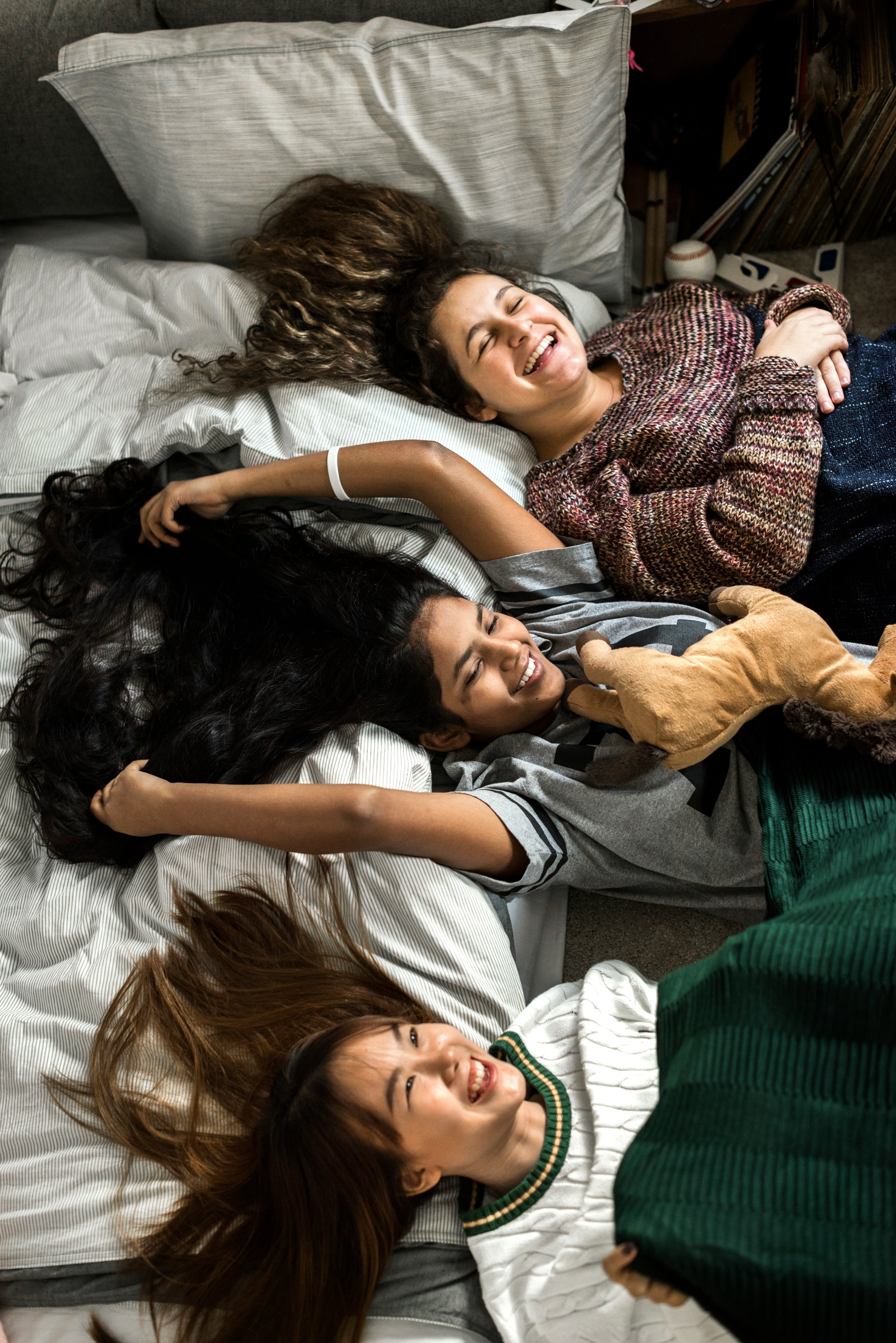 Three college friends lying side by side on a bed, laughing together in an overhead shot