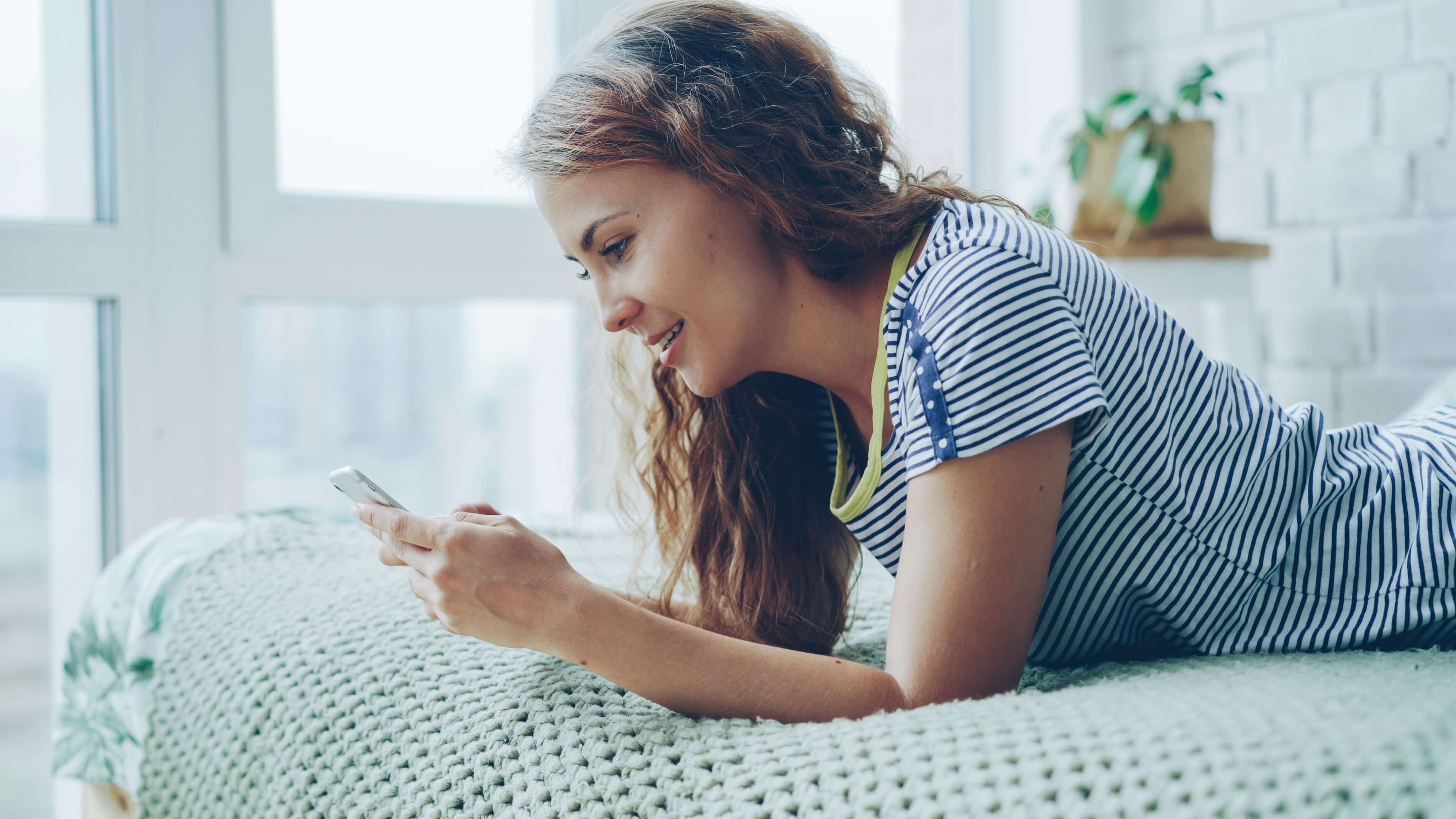 A young woman lying on a bed in a sunlit room, smiling as she reads a message on her phone