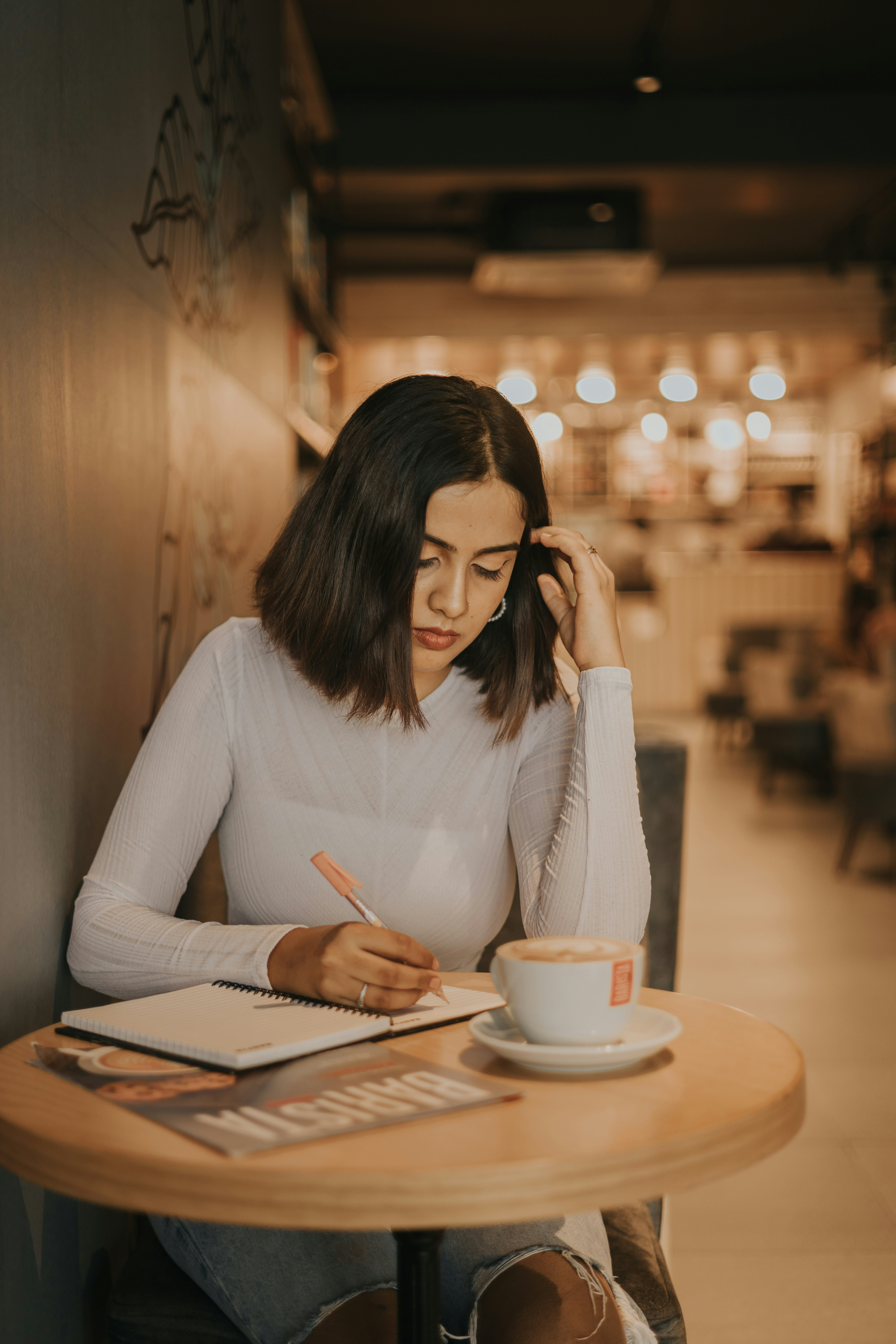 A young woman in a white top writing in a notebook at a small cafe table with a cappuccino, focused and at ease