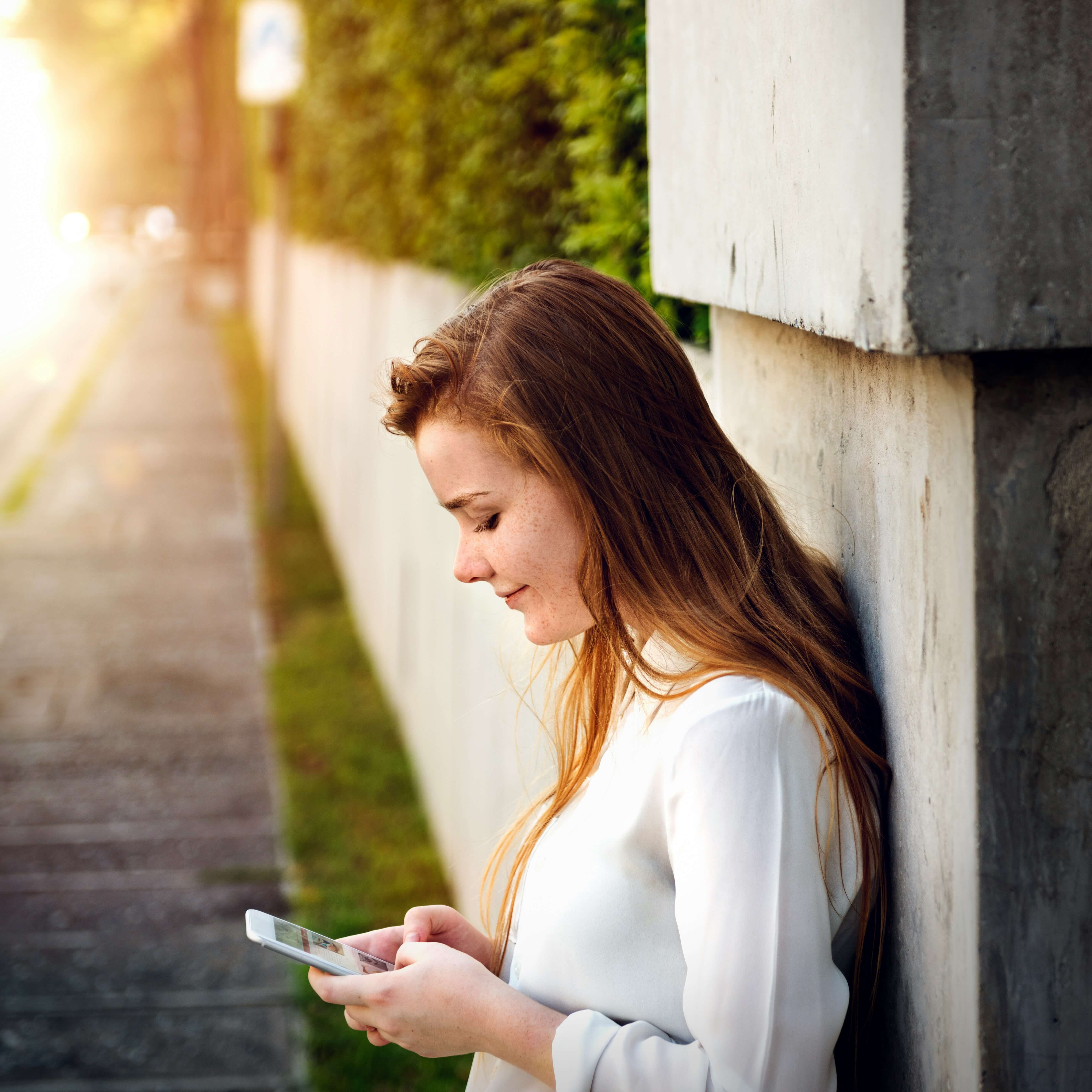 A young woman in a white top leaning against an outdoor wall, looking down at her phone in golden afternoon light