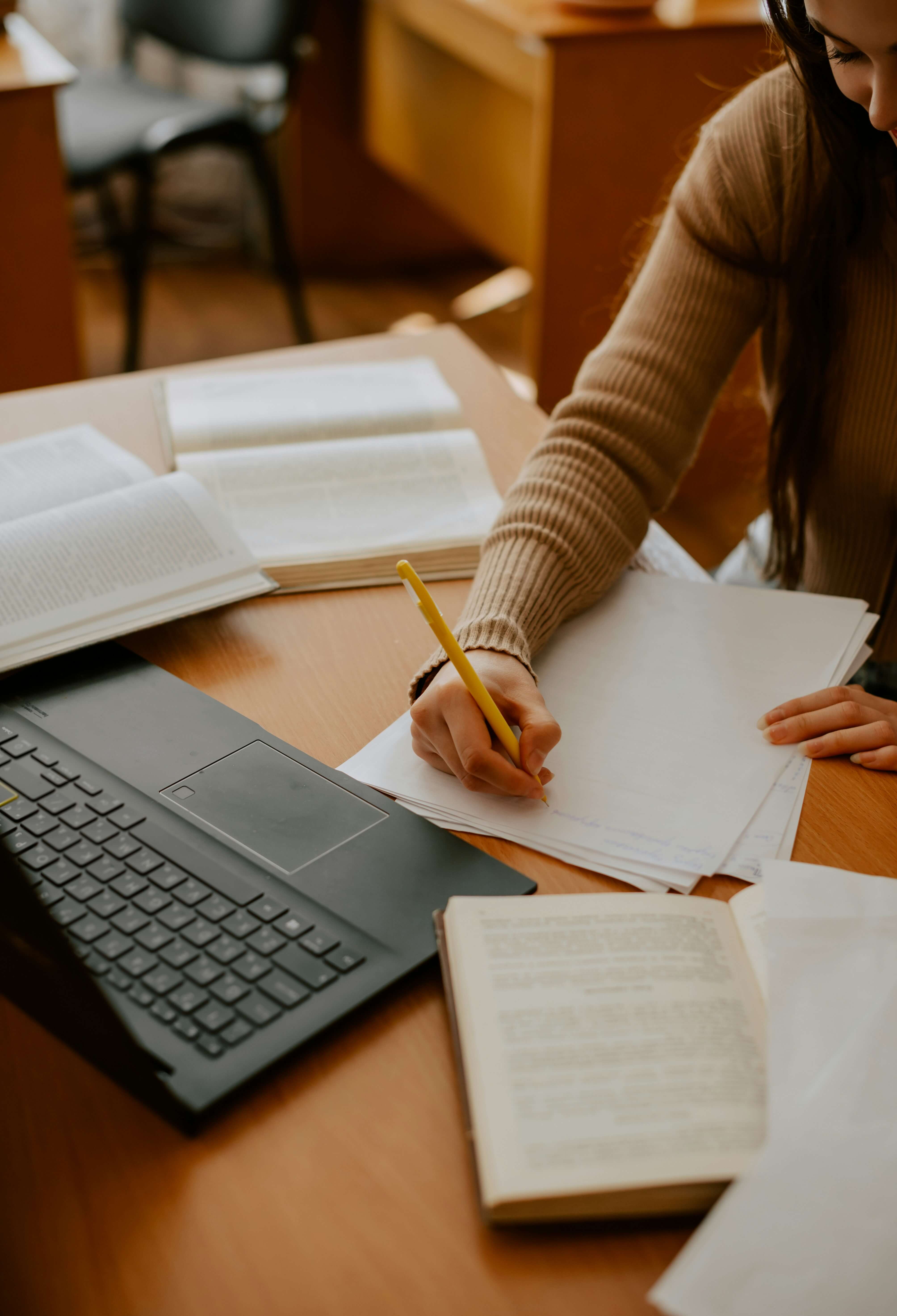 A young woman writing notes by hand at a desk surrounded by open books, papers, and a laptop